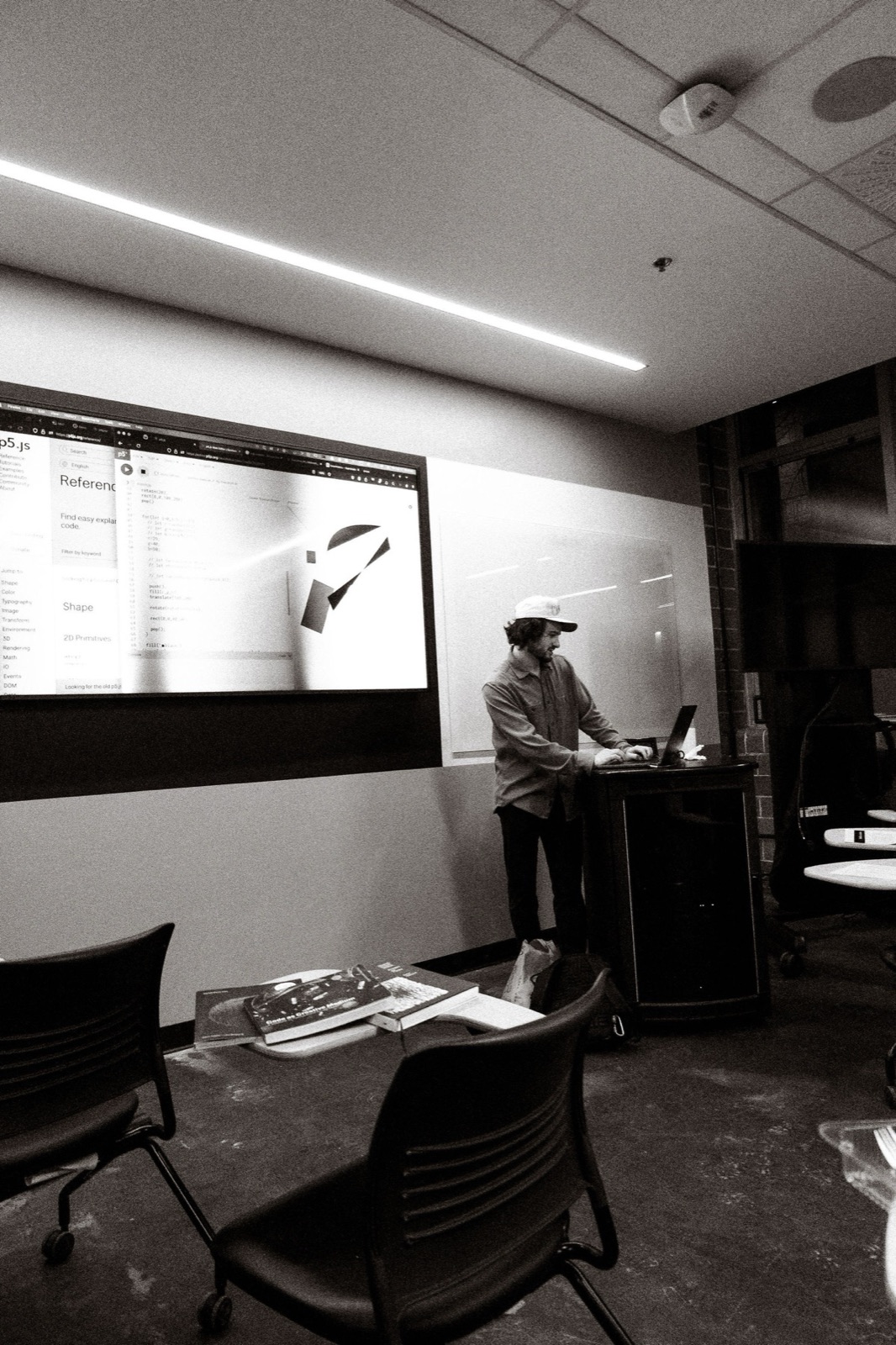 David Holcer presenting at the lectern, seen across the classroom in black and white